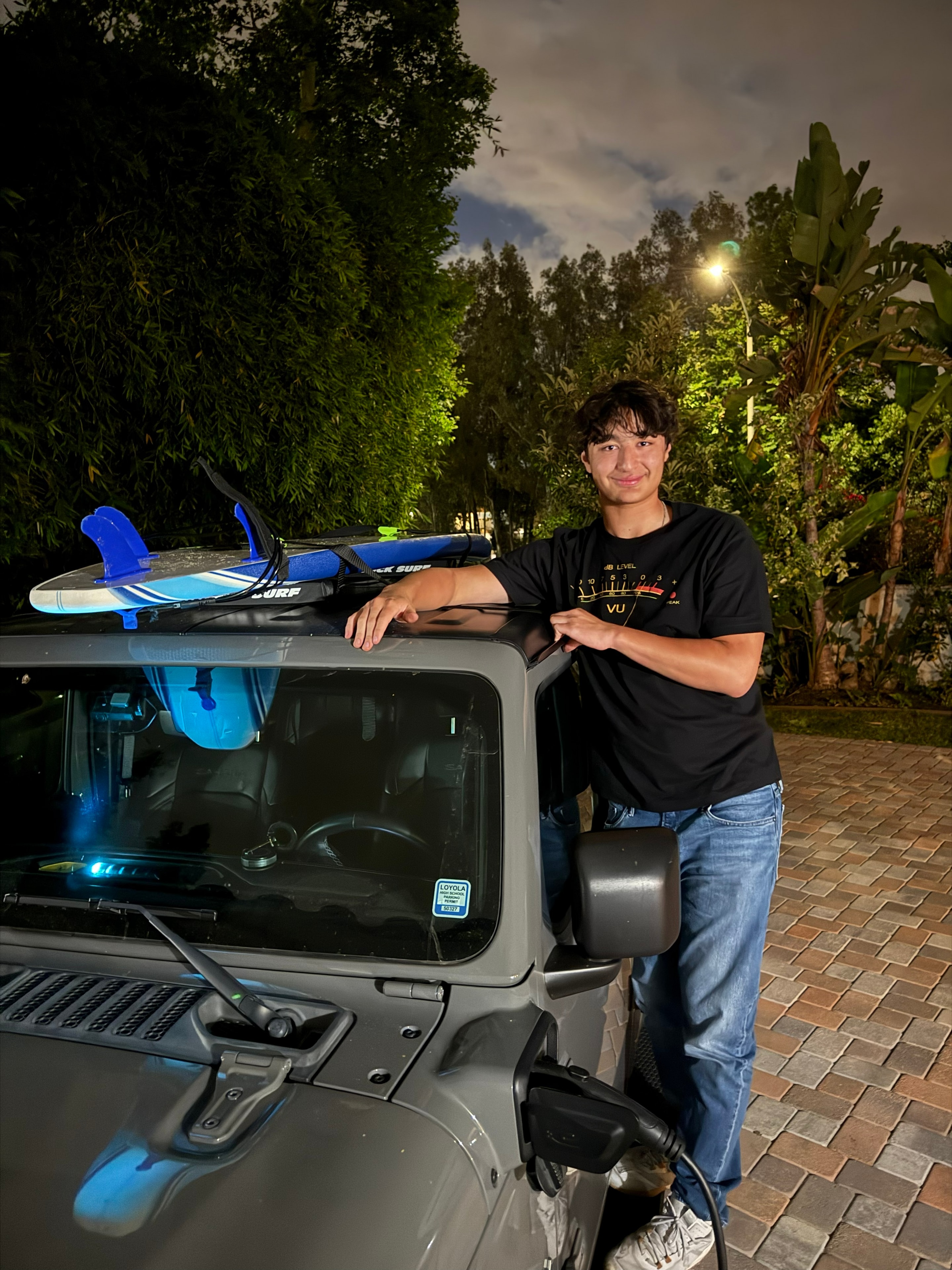 Teen boy leaning on Jeep with surfboard on roof at night